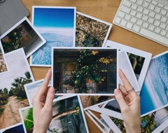 Op de afbeelding: Een verzameling afgedrukte foto's op een houten oppervlak, met afbeeldingen van landschappen en natuurscènes. Een persoon houdt een foto vast van een stenen gebouw met groen. Een laptop en een toetsenbord zijn ook zichtbaar.