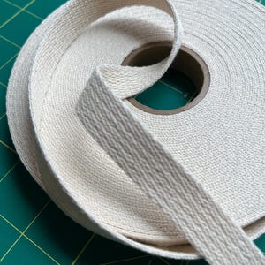 May include: A roll of off-white cotton twill tape featuring a distinct herringbone weave pattern. The fabric ribbon is wound tightly around a circular cardboard core, with one end of the tape extending outward from the center of the spool.