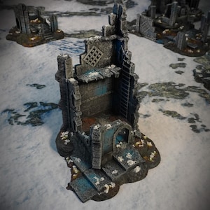 Puede incluir: Una miniatura de ruina de edificio de piedra desgastada con una pared parcialmente derrumbada, una escalera y una pequeña puerta. La estructura gris tiene detalles en azul y se asienta sobre una superficie nevada. Otras ruinas similares son visibles en el fondo, sugiriendo un juego de fantasía o de mesa.