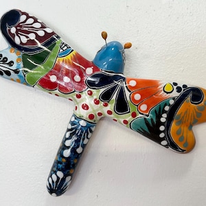 Puede incluir: Una colorida libélula de cerámica pintada a mano. Las alas presentan diseños en rojo, naranja, azul y verde. El cuerpo de la libélula es azul y tiene pequeñas antenas.