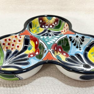 May include: A three-section ceramic serving dish featuring a vibrant hand-painted floral pattern in shades of yellow, red, green, and blue. The dish has a glossy finish with a thick black rim outlining each of the three rounded compartments.