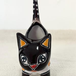Puede incluir: Figura de gato de cerámica con acabado negro brillante. El gato tiene orejas con puntas naranjas, ojos grandes y boca roja. Su cola está erguida y decorada con lunares blancos. El gato lleva una camisa blanca con un acento naranja.