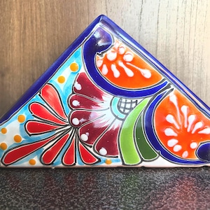 Puede incluir: Un servilletero triangular de cerámica con un vibrante diseño floral pintado a mano. El patrón incluye pétalos en tonos naranja, rojo y burdeos intenso sobre un fondo turquesa brillante, delineado en azul oscuro con puntos amarillos.