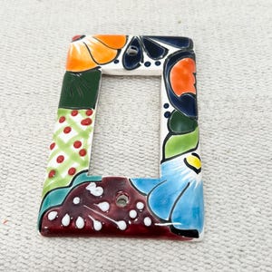 Puede incluir: Una placa de interruptor de luz rectangular de cerámica con un diseño floral pintado a mano. La placa presenta patrones florales en naranja, azul, verde y rojo. El diseño incluye una mezcla de colores sólidos y secciones estampadas, con dos orificios de montaje.