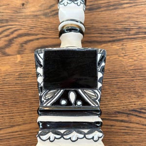 Puede incluir: Un decantador de cerámica con un esquema de color de alto contraste en blanco y negro. La botella tiene una base cuadrada con bordes moldeados en niveles y un cuello alto y delgado rematado con un tapón cuadrado. Patrones florales negros decoran la superficie blanca.