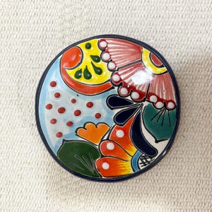 Puede incluir: Un posavasos redondo de cerámica con un diseño vibrante pintado a mano sobre un fondo azul claro, con puntos rojos y motivos florales estilizados en amarillo, naranja, rojo, verde y azul oscuro, bordeado por un círculo azul oscuro.