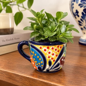 Puede incluir: Una pequeña taza de cerámica vibrante con un patrón pintado a mano estilo Talavera en azul profundo, amarillo brillante, naranja y rojo. La taza contiene una planta verde frondosa con follaje redondeado, creando un contraste con el asa azul.