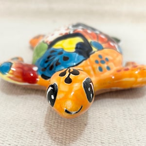 May include: A colorful ceramic turtle figurine with an orange head and flippers, and a shell decorated with blue, red, yellow, and green patterns. The turtle has a smiling face and black eyes. The figurine is a decorative item.