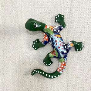 Puede incluir: Una figura de lagarto de cerámica con un cuerpo verde brillante y un diseño colorido en la espalda. El diseño incluye patrones florales y de puntos azules, naranjas y rojos. El lagarto tiene puntos blancos en las patas y la cola.