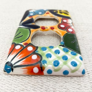 Puede incluir: Una cubierta de enchufe de cerámica pintada a mano con un vibrante diseño floral. La cubierta tiene forma rectangular con dos aberturas y está adornada con patrones naranjas, rojos, verdes, azules y blancos. El diseño incluye flores estilizadas y formas geométricas.