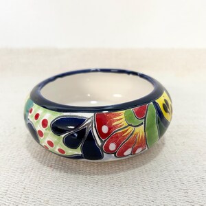 Puede incluir: Un cuenco pequeño y redondo de cerámica con un diseño floral vibrante. El cuenco tiene un borde azul oscuro y un interior blanco. El exterior está adornado con flores coloridas, hojas y lunares en rojo, verde, amarillo y azul. Una pieza decorativa.