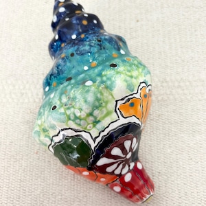 Puede incluir: Una escultura de concha marina de cerámica pintada a mano. La concha presenta una vibrante paleta de colores de azules, verdes, naranjas y rojos, con acentos de puntos blancos y negros. El diseño incluye un motivo de mariposa.