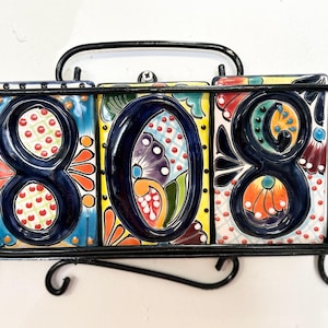 May include: A black metal wall-mounted frame holding three rectangular ceramic tiles, each featuring a raised dark blue number 8, 0, and 8. The tiles are decorated with colorful hand-painted floral and geometric patterns in orange, green, and yellow.