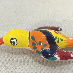 Puede incluir: Figura de pájaro de cerámica colorida. El pájaro tiene cabeza amarilla, cuerpo naranja y pico rojo. Las alas están decoradas con patrones azules, naranjas y blancos. La figura mide aproximadamente 13 cm de largo.