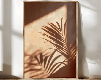 Pode incluir: Uma impressão de arte de parede retangular numa moldura de madeira clara com um fundo quente de cor terracota e a sombra escura e nítida de folhas de palmeira projetada na superfície, criando um efeito visual de alto contraste com tons claros e escuros.