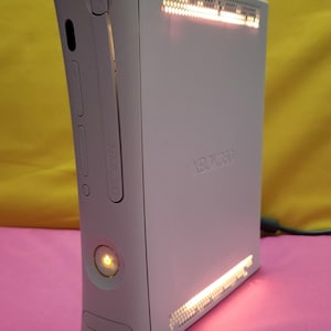 May include: A white Xbox 360 console with the Xbox 360 logo embossed on the side. The console has a power button and a disc tray. The console is lit up with orange and red lights. The console is on a pink surface.
