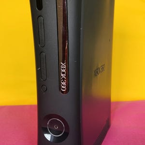 May include: Black Xbox 360 console with a silver trim and the Xbox 360 logo. The console has a power button and a disc tray. The background is a yellow and pink gradient.