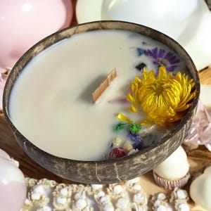 Peut inclure: Une bougie dans une noix de coco avec de la cire blanche crémeuse, une mèche en bois et des fleurs séchées. La bougie présente une grande fleur jaune et d'autres fleurs séchées colorées. La noix de coco est marron.