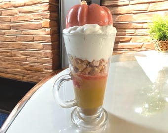 May include: A layered dessert in a clear glass with a handle, topped with a pink pumpkin. The layers include a yellow liquid, a reddish layer, a crumbly layer, and a white, whipped cream-like layer. The glass sits on a white surface.