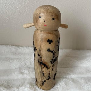 May include: A wooden doll with a rounded head, small arms, and a cylindrical body. The doll's face is hand-painted with black eyes and a red mouth. The body features dark, organic patterns. The doll is approximately 8 inches tall.