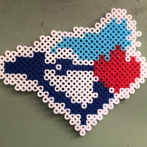 Puede incluir: Un mosaico de cuentas blancas, azules y rojas en forma de corazón. El mosaico está hecho de pequeñas cuentas redondas que están dispuestas en un patrón.
