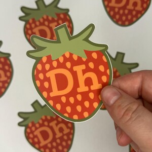 May include: A strawberry-shaped sticker with a green stem and leaves. The red body of the strawberry is dotted with orange seeds and features the orange letters "Dh". Several similar stickers are visible in the background.