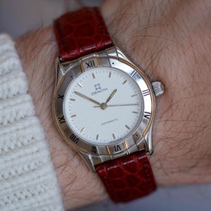 May include: A Zenith automatic wristwatch with a silver and gold-toned case and a white dial. The watch features Roman numeral hour markers and gold hands. It has a red leather strap.
