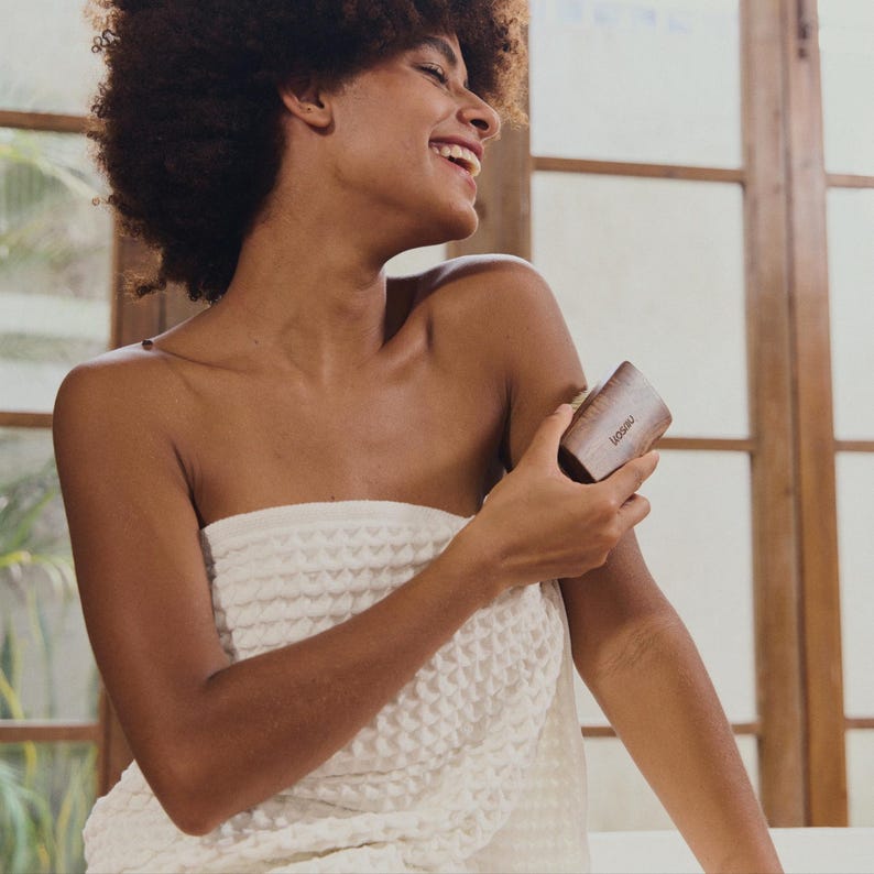 May include: A woman wrapped in a white towel smiles while using a wooden body massager. The massager is rectangular with rounded edges and the word "leosaz" is printed on the side. The background includes a window and a white tiled surface.