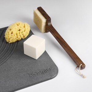 May include: A spa set featuring a wooden handled brush with natural bristles, a yellow sponge, a white cube of soap, and a gray textured tray with the word "kosmu" embossed on it. The brush has a loop for hanging.