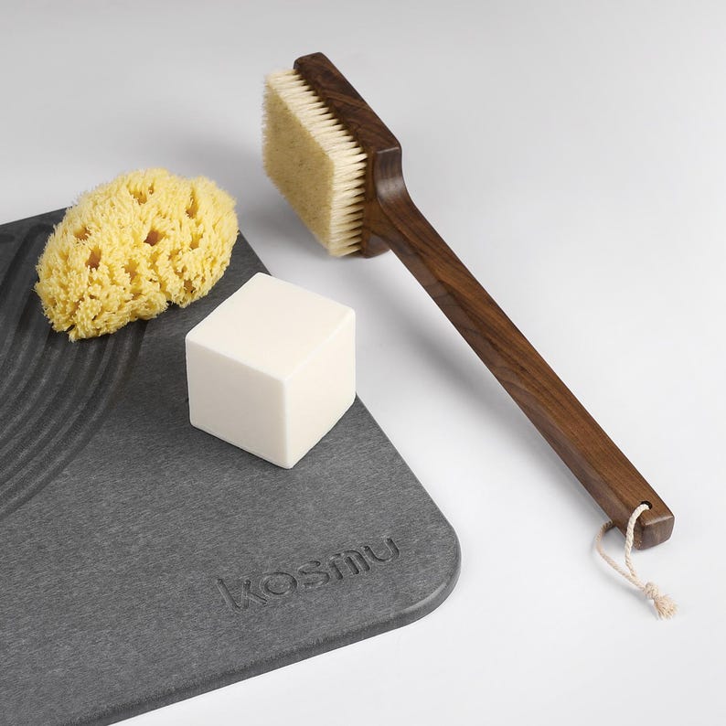 May include: A spa set featuring a wooden handled brush with natural bristles, a yellow sponge, a white cube of soap, and a gray textured tray with the word "kosmu" embossed on it. The brush has a loop for hanging.
