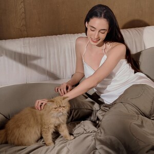 May include: A woman in a white satin tank top sits on a bed with a brown and white cat. The bed has a brown quilted cover and white pillows.