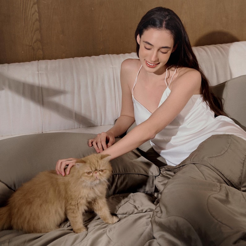 May include: A woman in a white satin tank top sits on a bed with a brown and white cat. The bed has a brown quilted cover and white pillows.