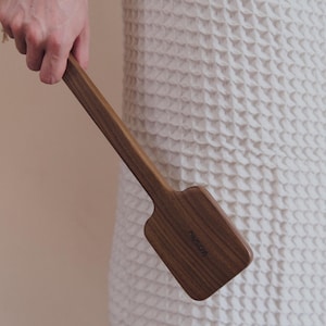 May include: A wooden spatula with a wide, rectangular head and a long, slender handle. The spatula is made of a dark brown wood and has the word "kosmu" etched on the head. The spatula is held against a white, textured background.