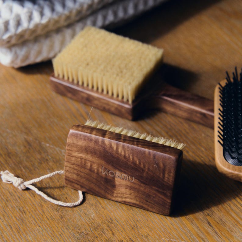 May include: Wooden body brushes with natural bristles. One brush has a loop of white string attached. The brushes are made of dark wood and have a light-colored bristle section. The word "kosmu" is printed on one brush.