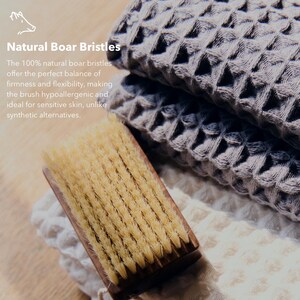 May include: A close-up of a wooden-handled brush with natural boar bristles, alongside folded gray waffle-weave towels. The text "Natural Boar Bristles" is visible. The brush is designed for sensitive skin.