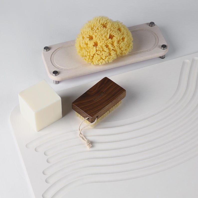 May include: A natural sea sponge, wooden brush, and bar of soap are displayed on a white surface. The brush has a dark wood handle and light bristles. A light-colored wooden tray holds the yellow sponge. The soap is a white cube.