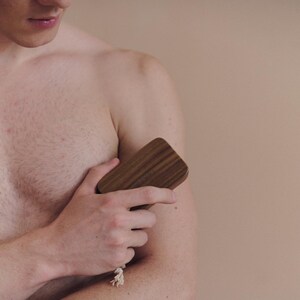 May include: A wooden body brush with a rectangular shape and rounded edges. The brush is made of dark brown wood and has a light-colored rope attached to it. The brush is being held against a person's arm.