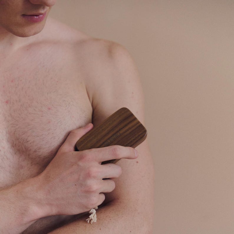 May include: A wooden body brush with a rectangular shape and rounded edges. The brush is made of dark brown wood and has a light-colored rope attached to it. The brush is being held against a person's arm.