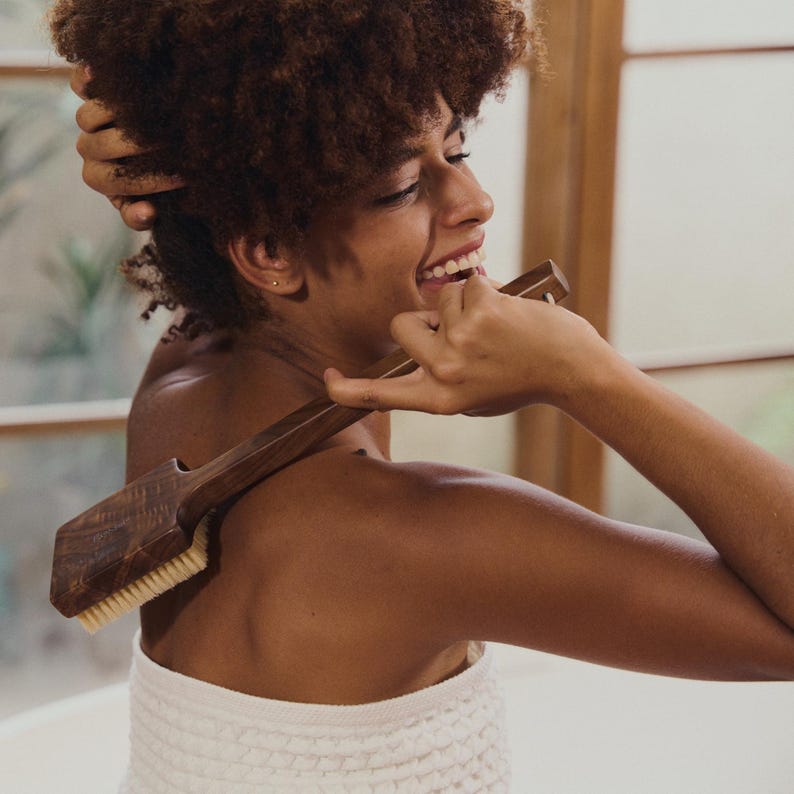May include: A wooden back brush with a long handle and soft bristles. The brush is dark brown and has a rectangular head. The person is smiling and holding the brush.