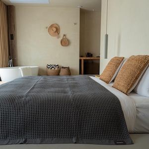 Peut inclure: Une couverture grise foncée en nid d'abeille recouvre un lit avec des draps blancs et deux oreillers décoratifs tissés. La pièce a une palette de couleurs neutres, avec une baignoire, des paniers et un chapeau au mur. La couverture est un élément clé de cette chambre.
