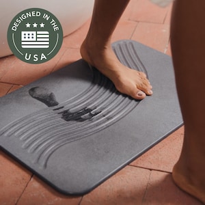 May include: A gray rectangular bath mat with a wave-like textured design. The mat has a wet footprint on it. The mat has the text "DESIGNED IN THE USA" in a circular emblem.