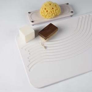 May include: A white rectangular bath mat with wave-like grooves, a natural sea sponge on a wooden tray, a wooden-handled brush, and a white bar of soap. The items are arranged on a white surface, creating a minimalist aesthetic.