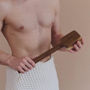 May include: A wooden body brush with a long handle. The brush head is rectangular with soft bristles. The handle is a dark brown color, and the brush is held by a person wrapped in a white towel.