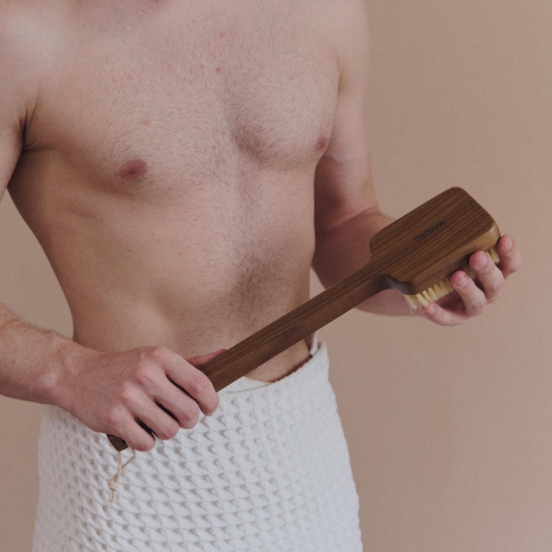 May include: A wooden body brush with a long handle. The brush head is rectangular with soft bristles. The handle is a dark brown color, and the brush is held by a person wrapped in a white towel.