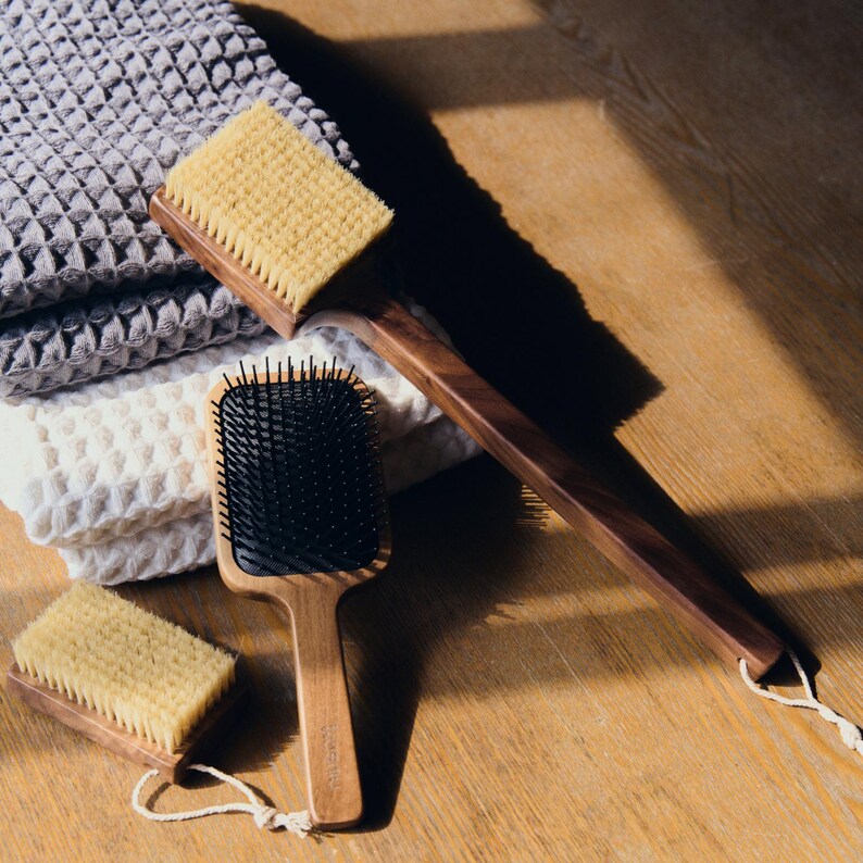 May include: A collection of wooden bath and hair brushes with natural bristles and a black bristle hairbrush. The brushes are displayed on a wooden surface next to folded gray and white waffle-weave towels. The long-handled brush is ideal for back scrubbing.