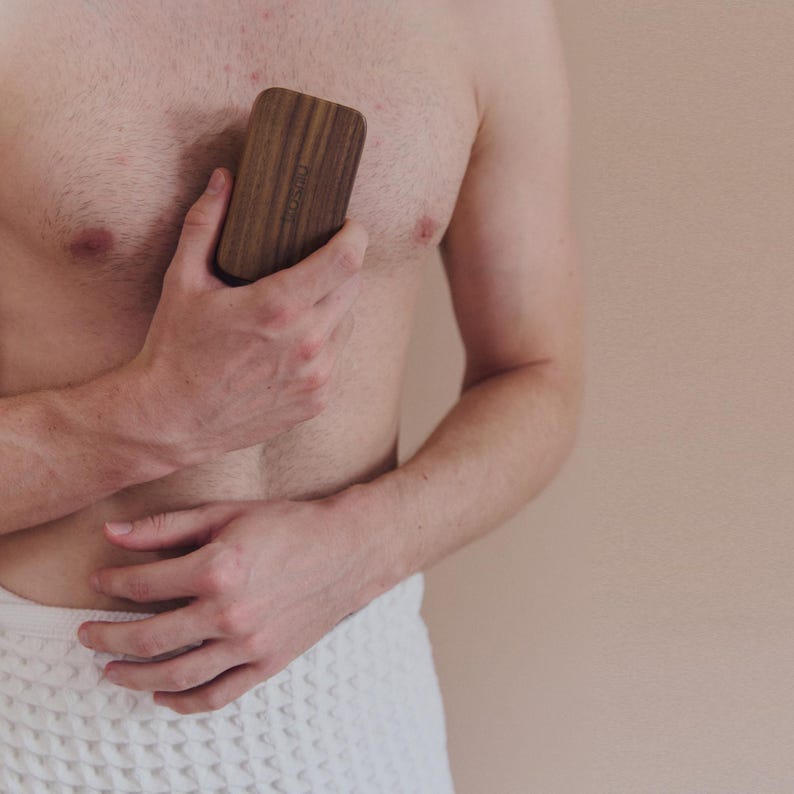May include: A rectangular wooden massage tool with rounded edges is held against a person's chest. The tool has a natural wood grain finish and a subtle, engraved design. The person is wearing a white towel.