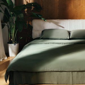 Puede incluir: Una cama verde con una cabecera blanca y dos almohadas verdes. La cama está hecha con una funda nórdica verde y una falda de cama verde. Una gran planta verde está en una maceta en el suelo junto a la cama.
