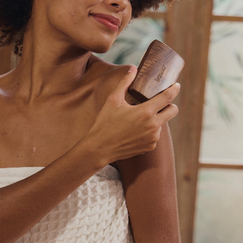 May include: A wooden massage tool with the word "losani" printed on it. The tool is rectangular with rounded edges and is being held by a person. The person is wrapped in a white towel.