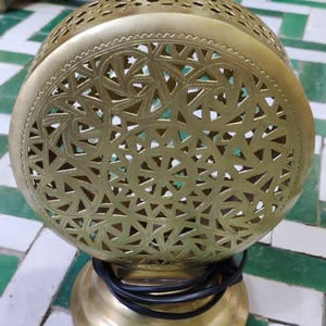 May include: A gold-colored, spherical lamp with intricate geometric cutouts. The lamp sits on a round base and has a black power cord. Other similar lamps are visible in the background. The lamp is likely for home decor.