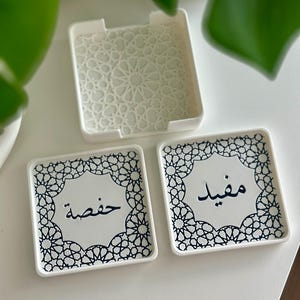 May include: Set of three white ceramic coasters with black geometric designs. Two coasters feature Arabic calligraphy, one says "Mehfil" and the other says "Khujista".